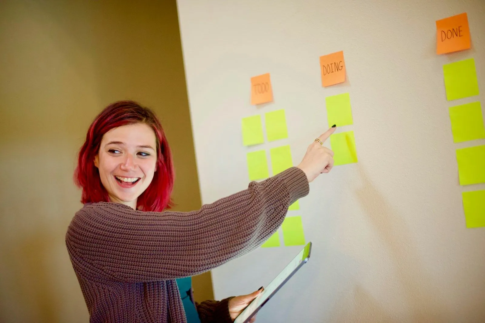 A woman with red hair smiles as she points to sticky notes on a wall.