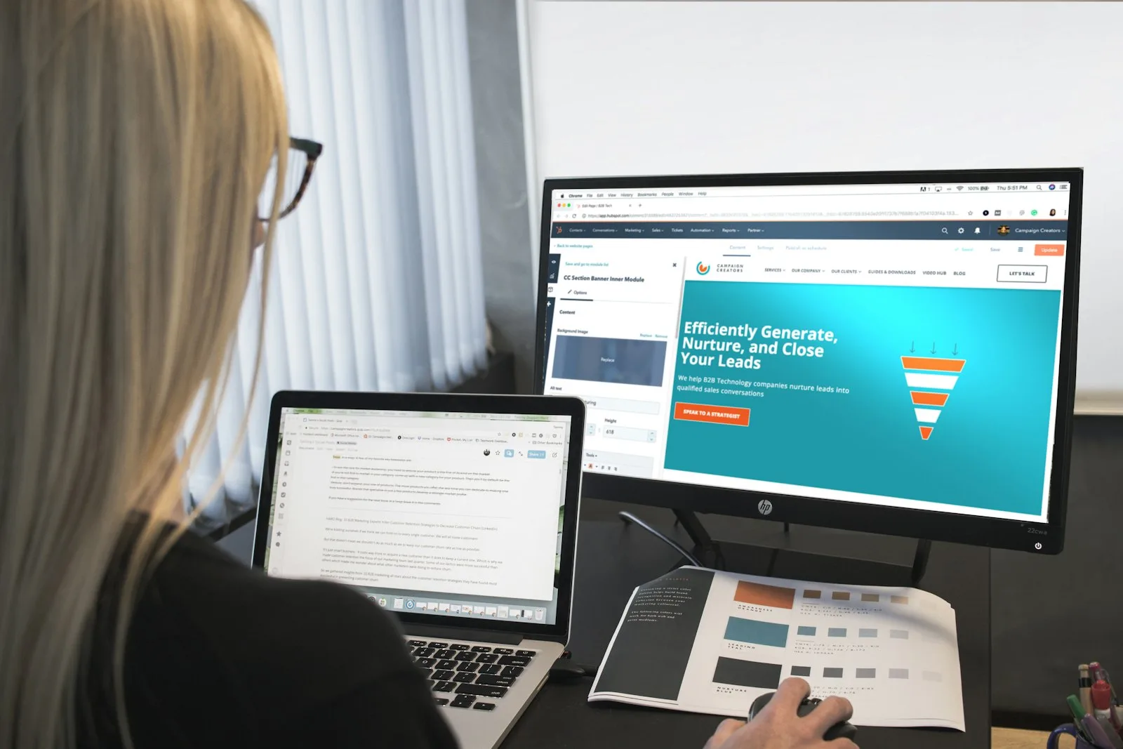 A UI/UX designer working on a web page layout at her desk, colour swatches printed beside her laptop.