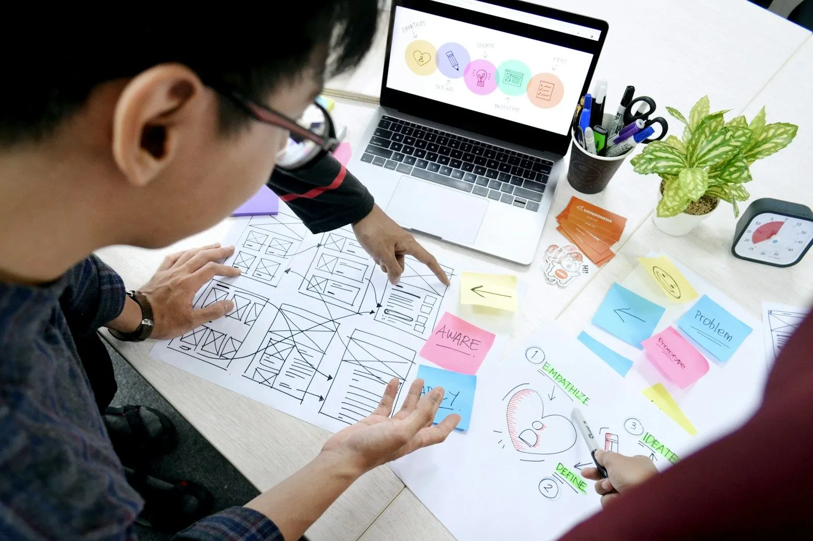 A product designer in glasses sketches UI/UX wireframes beside sticky notes and a laptop.