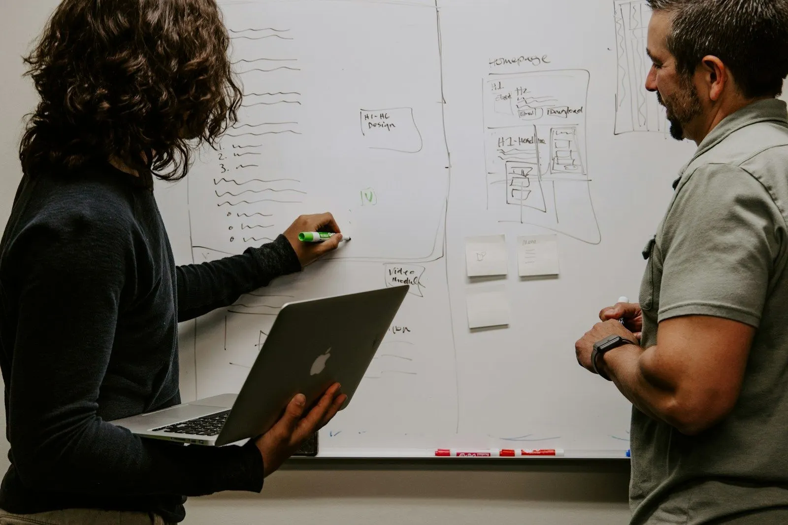 A data business analyst working through a process diagram on a whiteboard, a colleague holding a laptop.