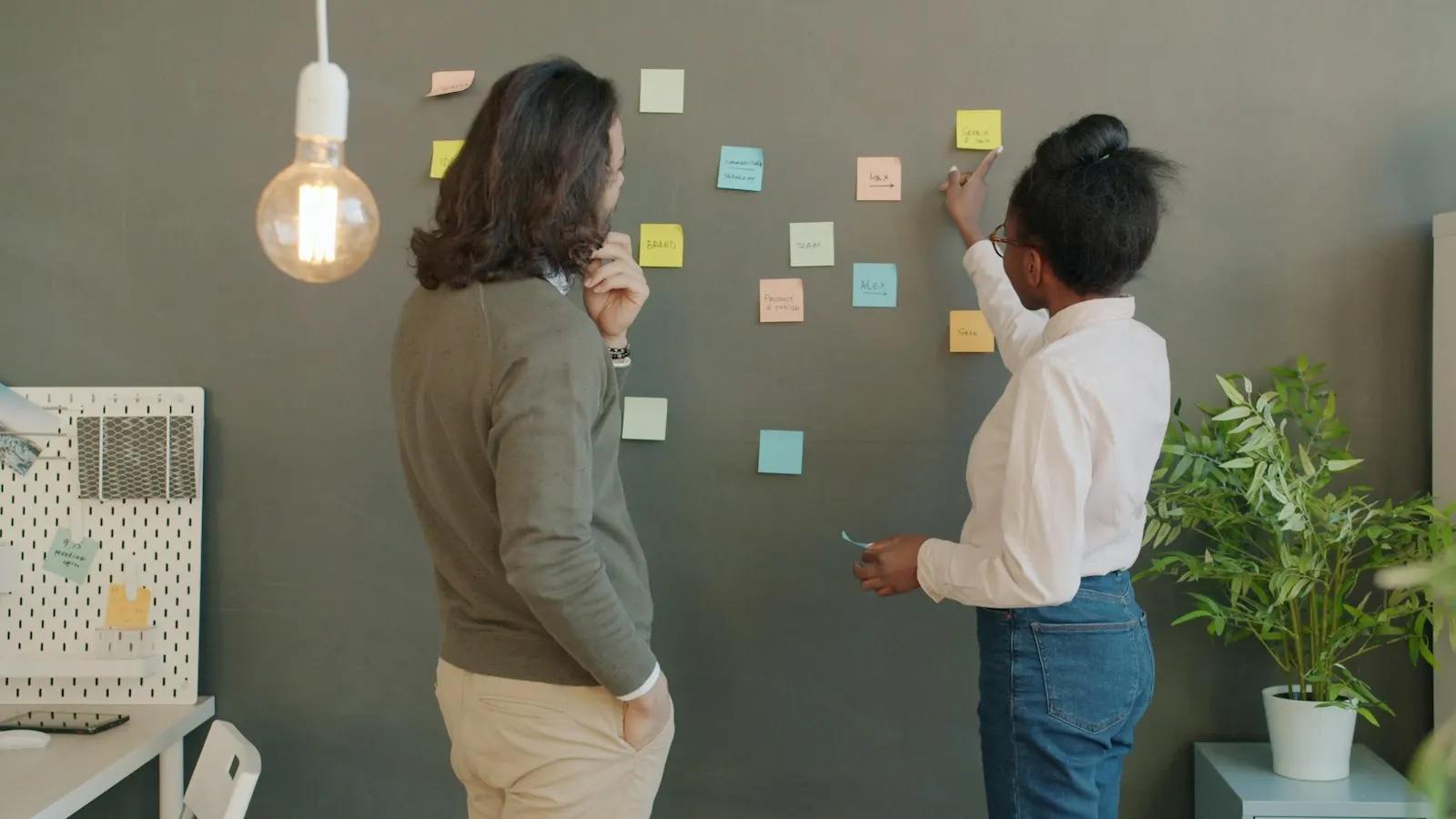 Two business analysts discussing a wall of sticky notes in a bright office.