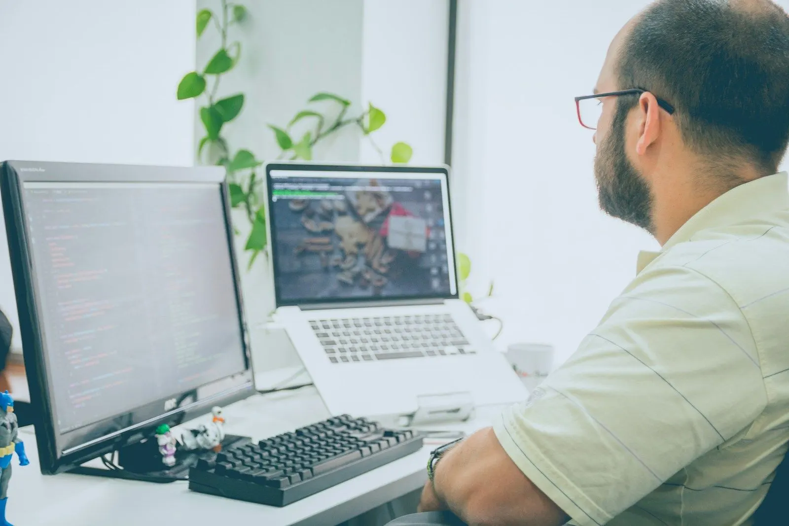 A React developer in glasses working on code across a monitor and a laptop by a window.