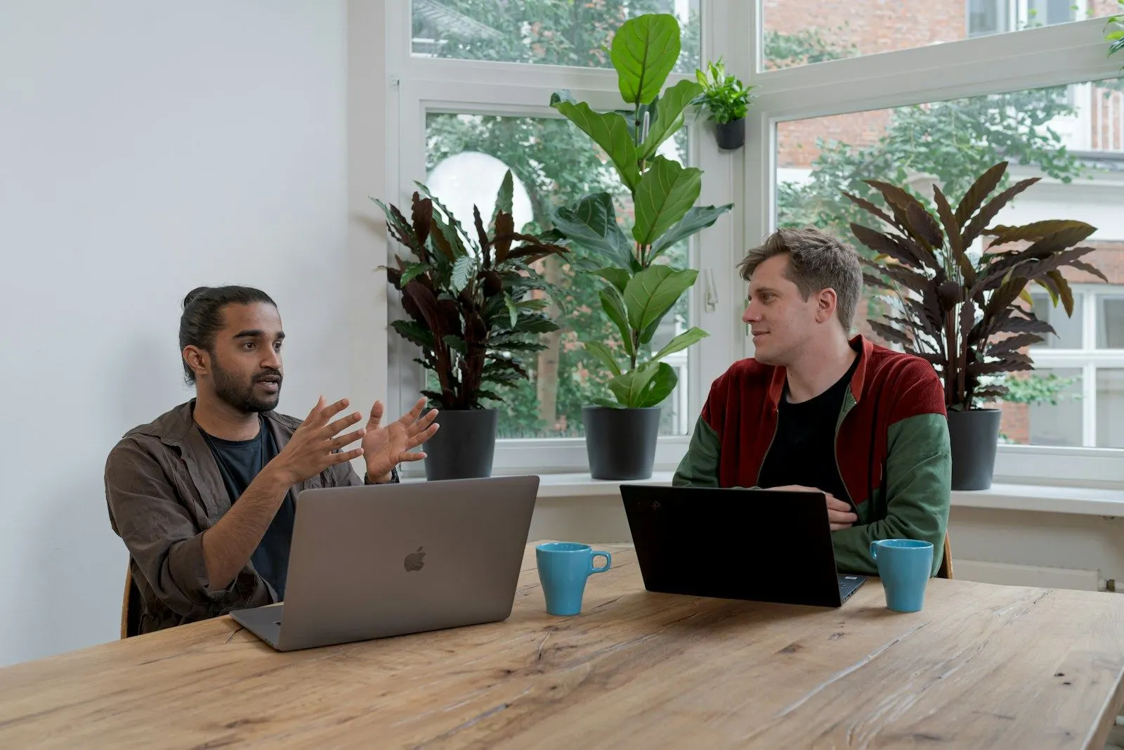 Two colleagues discussing how to hire a Node.js developer at a table with laptops.