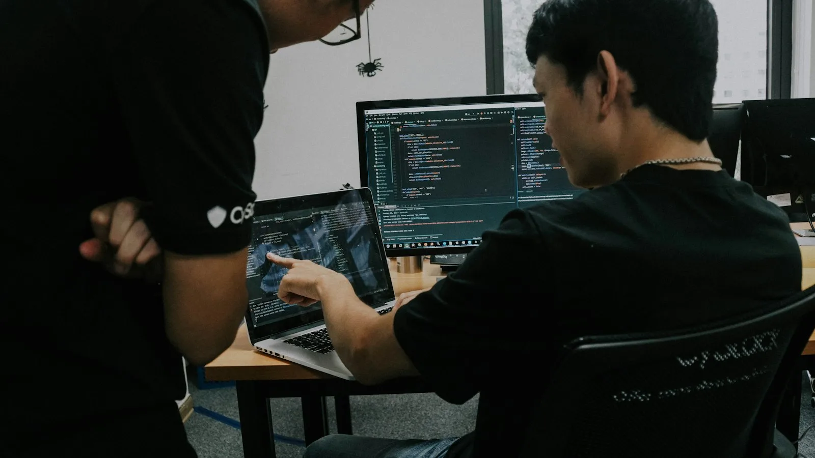 A Node.js developer pointing out a line of code on a laptop to a colleague at his desk.