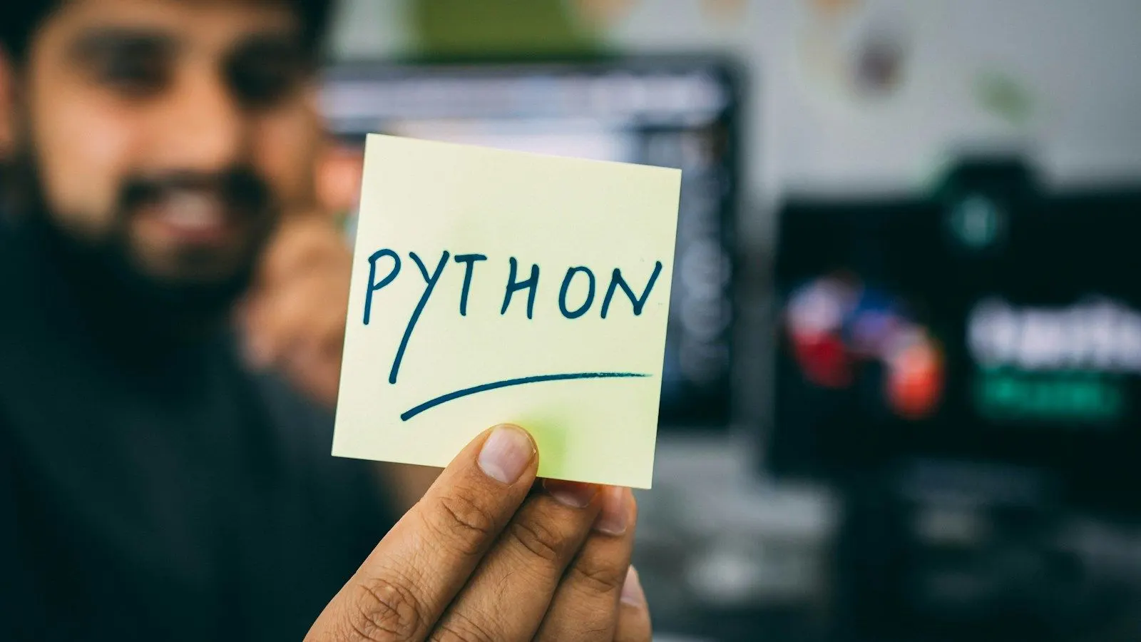 A Python backend architect holding up a sticky note that reads “Python” in front of his screens.