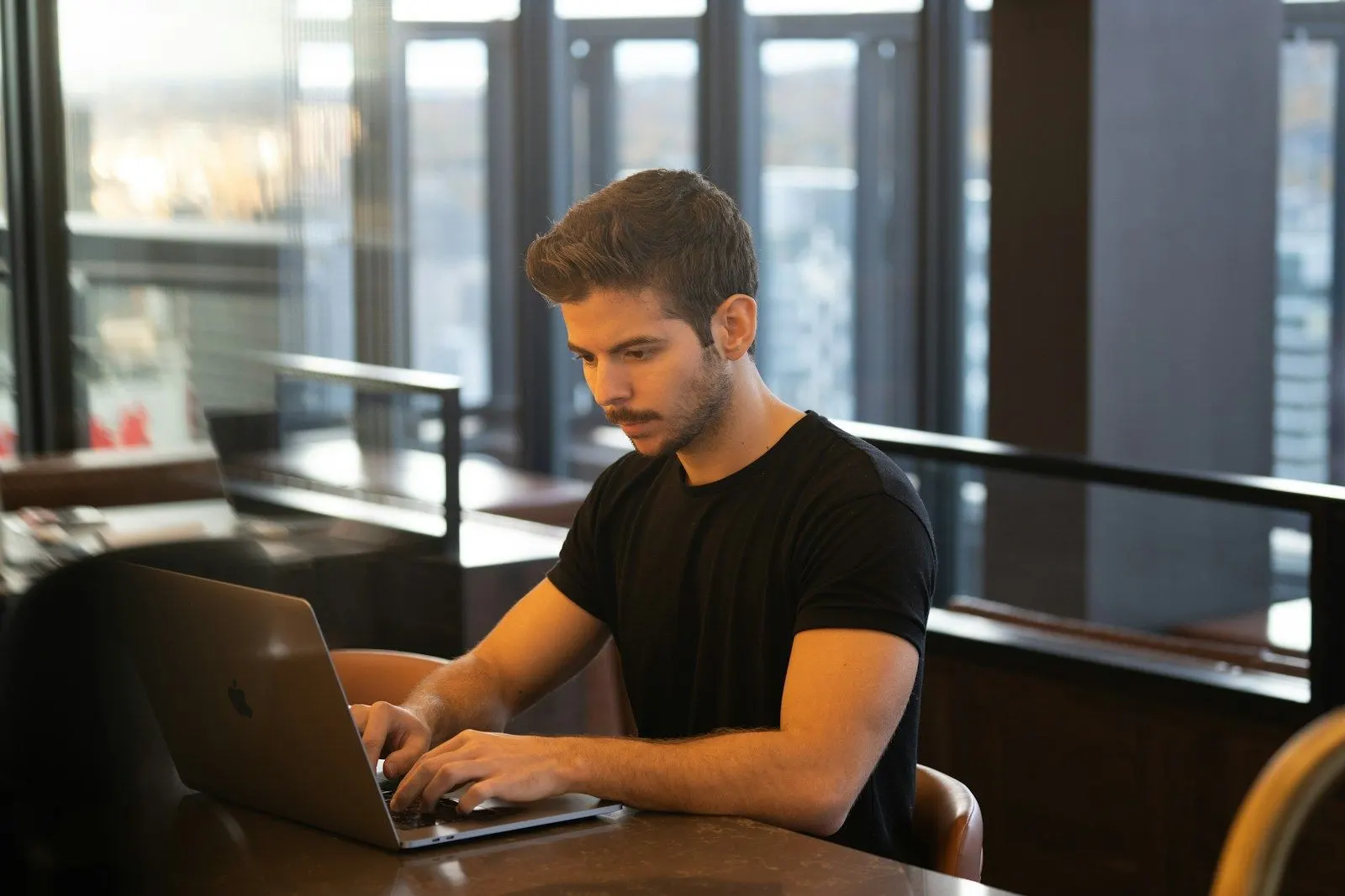 A .NET specialist in a black t-shirt working on a laptop at a long table in a bright office.