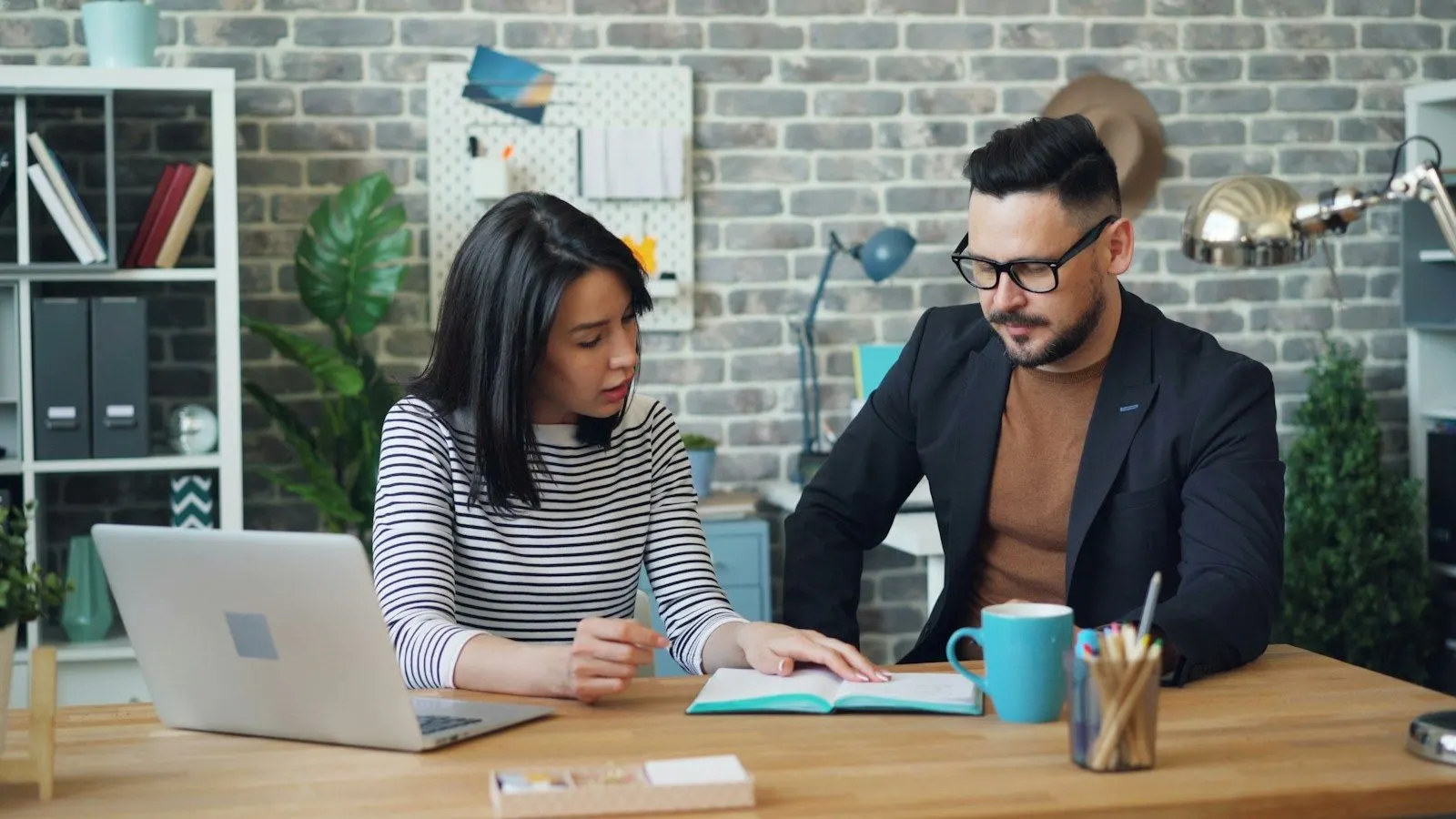 Two colleagues discussing how to hire a Java developer at a desk with a laptop in a brick-walled office.