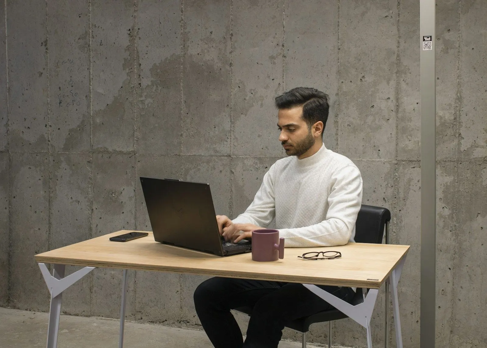 A Java developer in a white sweater working on a laptop at a simple desk against a concrete wall.
