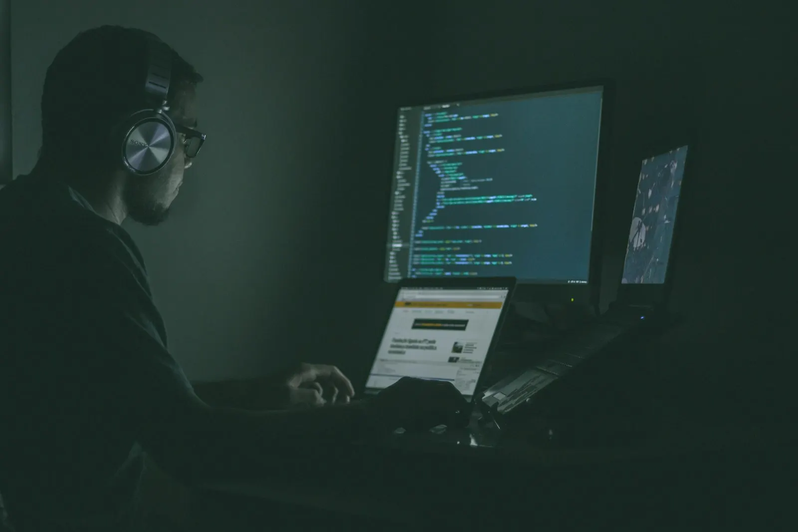 A PHP developer in headphones writing code across his monitors in a dim room.
