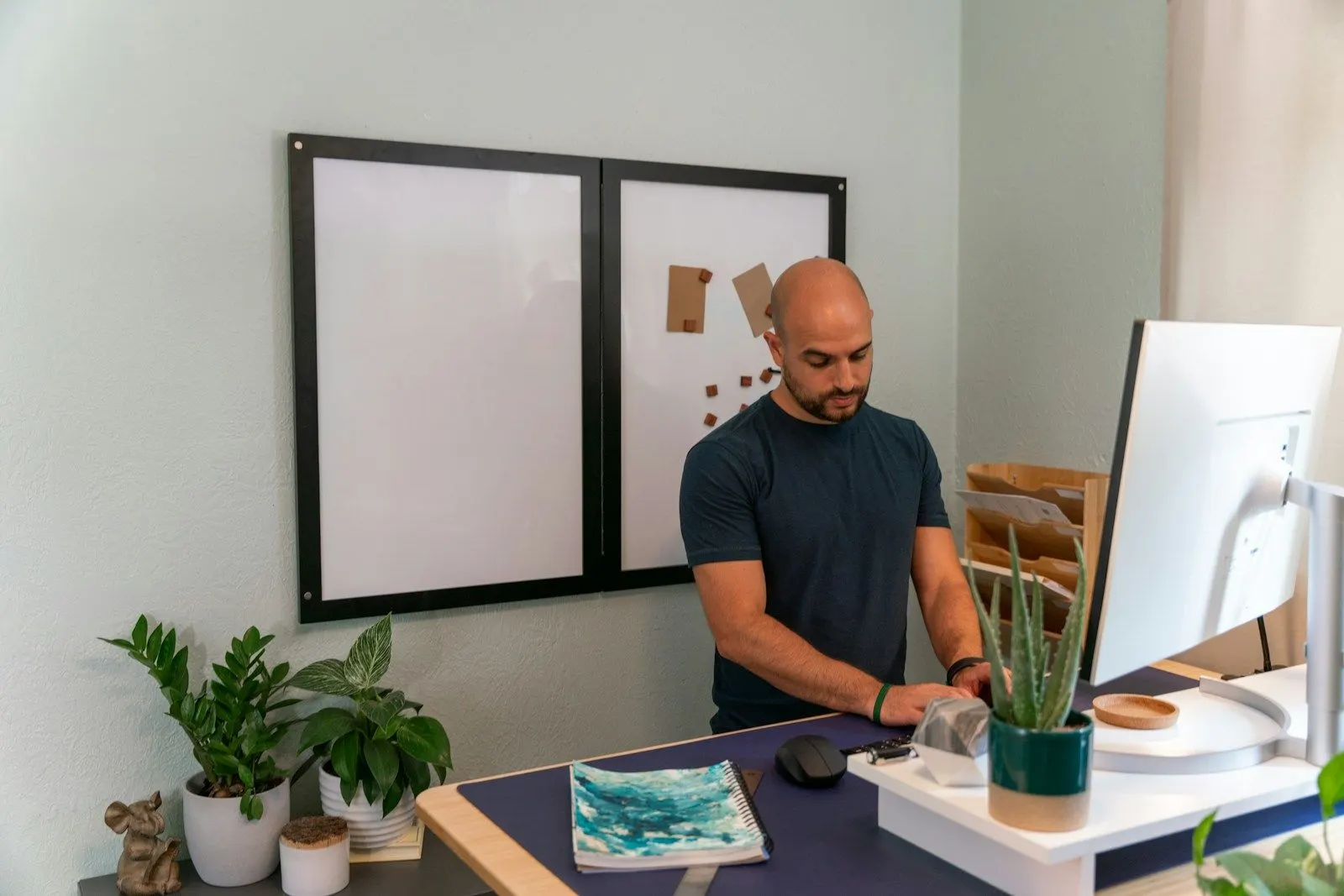 A PHP architect in a black t-shirt working at a standing desk with an all-in-one computer in a plant-filled studio.