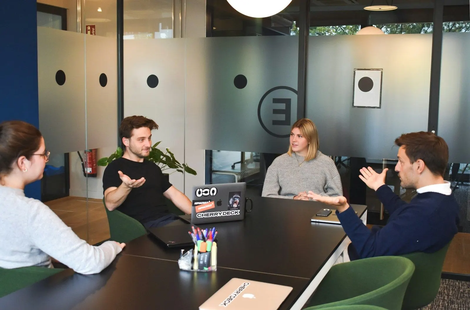 A small team talking through how to hire a PHP developer around laptops in a glass-walled meeting room.