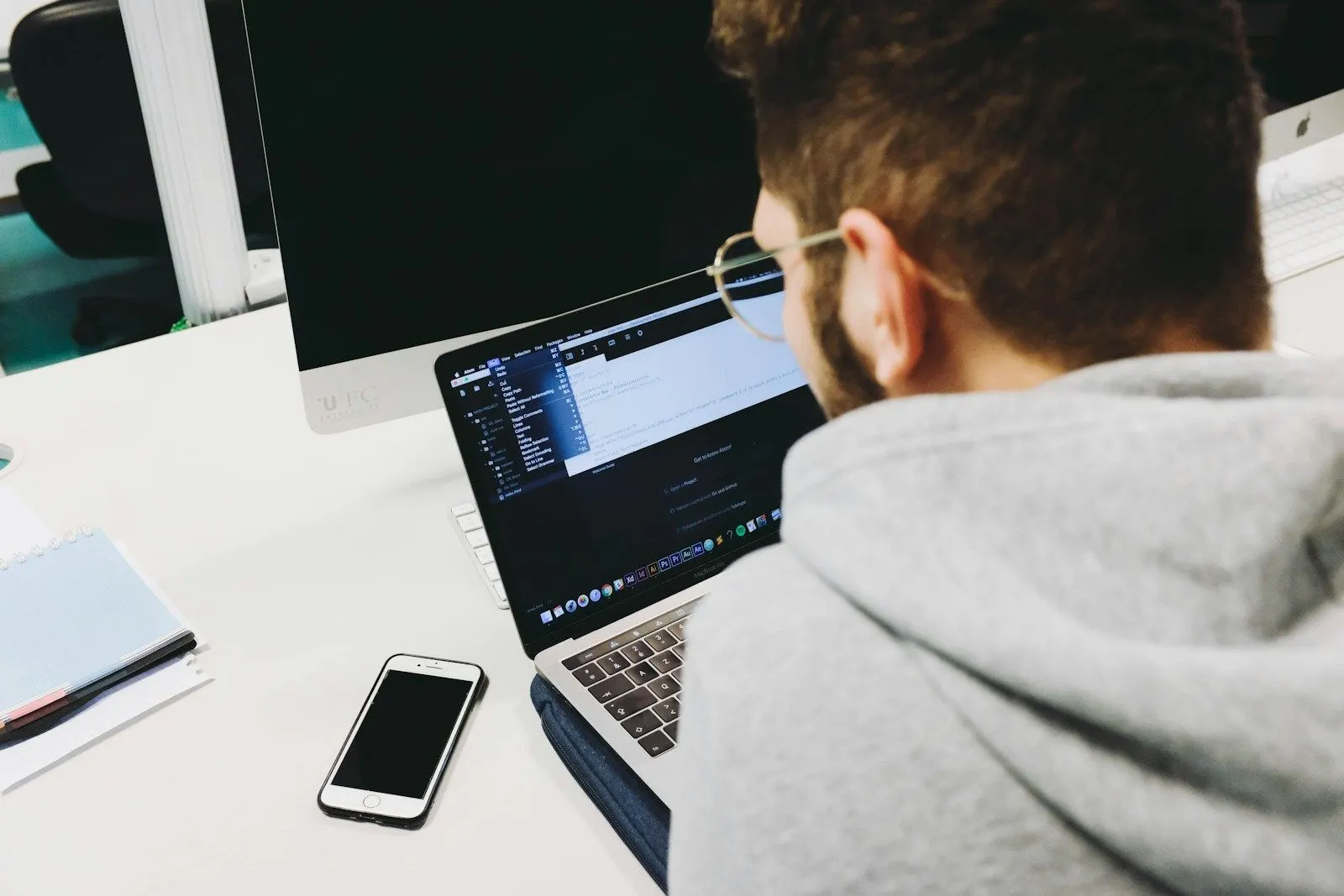 A PHP developer in a grey hoodie working in a code editor on his laptop, phone beside him.