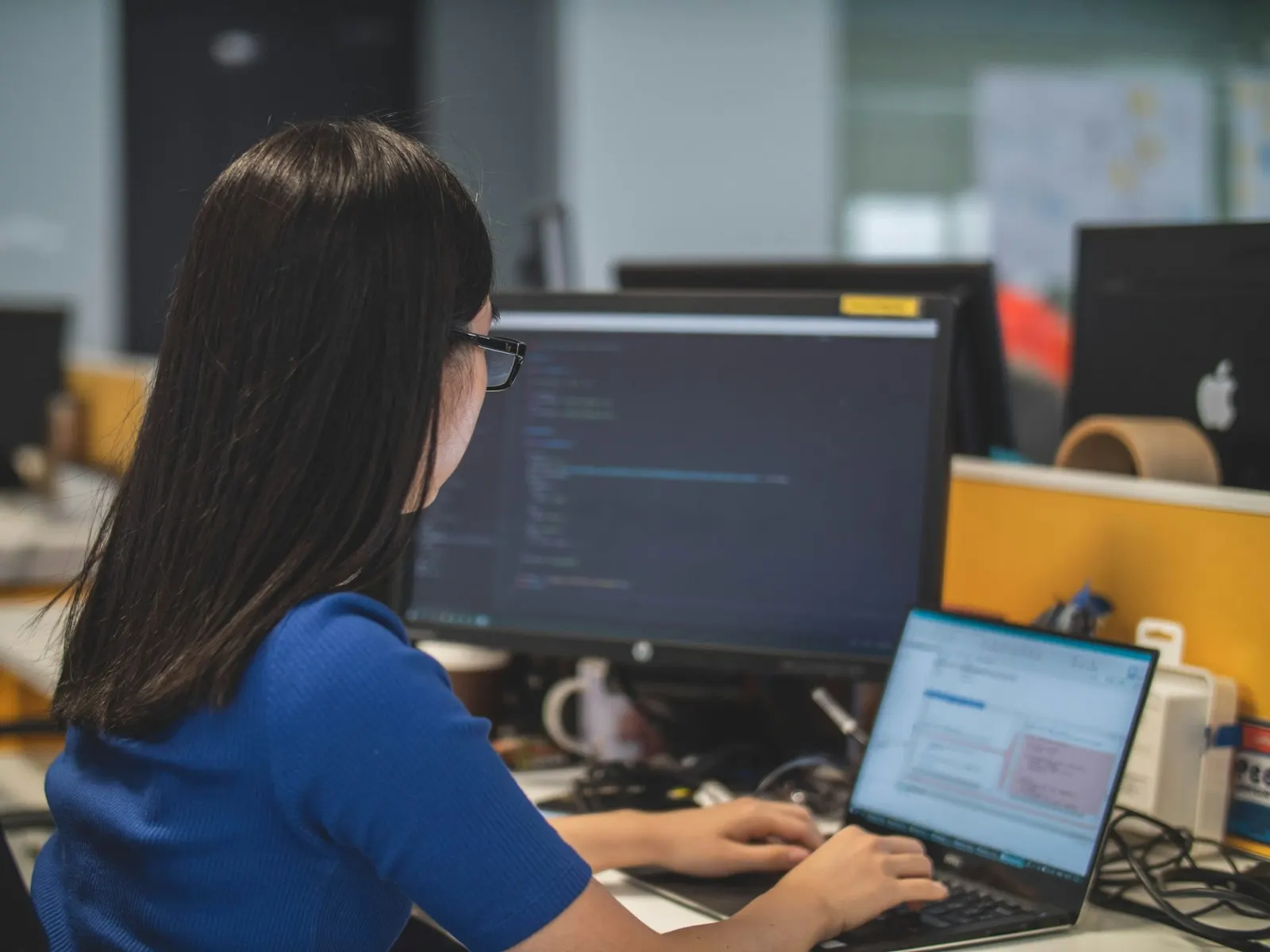 A React Native developer writing code at a desk with two screens in a busy studio.