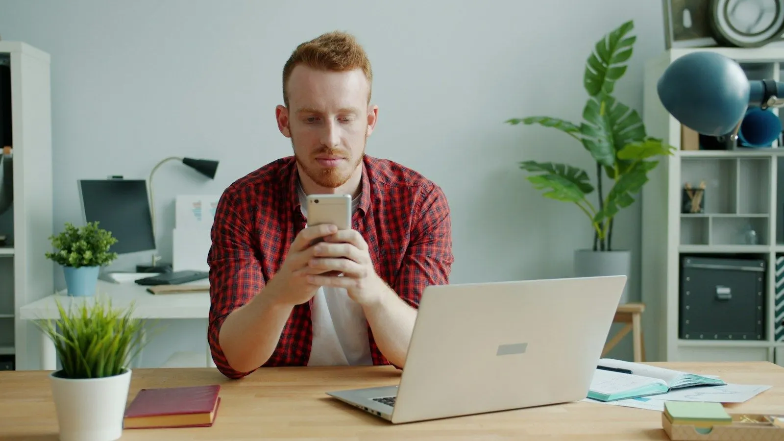 A React Native architect in a red plaid shirt testing an app on his phone at his laptop desk.