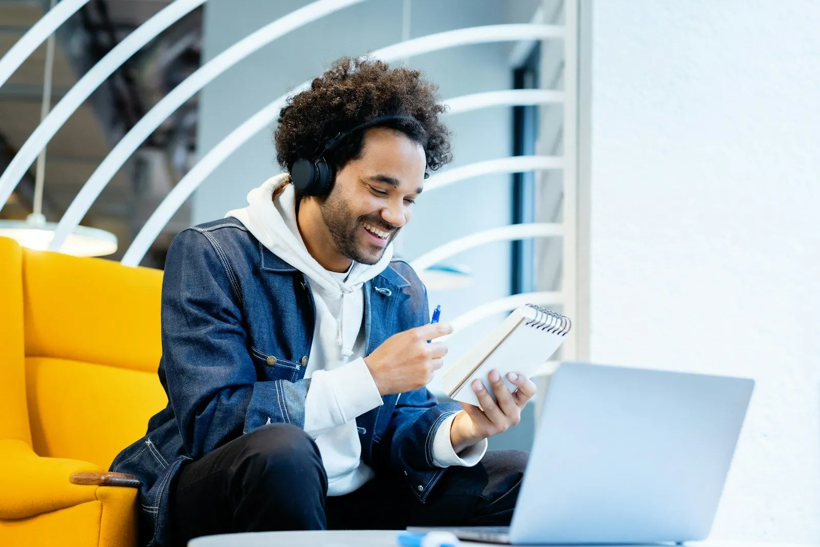 A React Native developer in a denim jacket and headphones smiling over his notes beside a laptop.