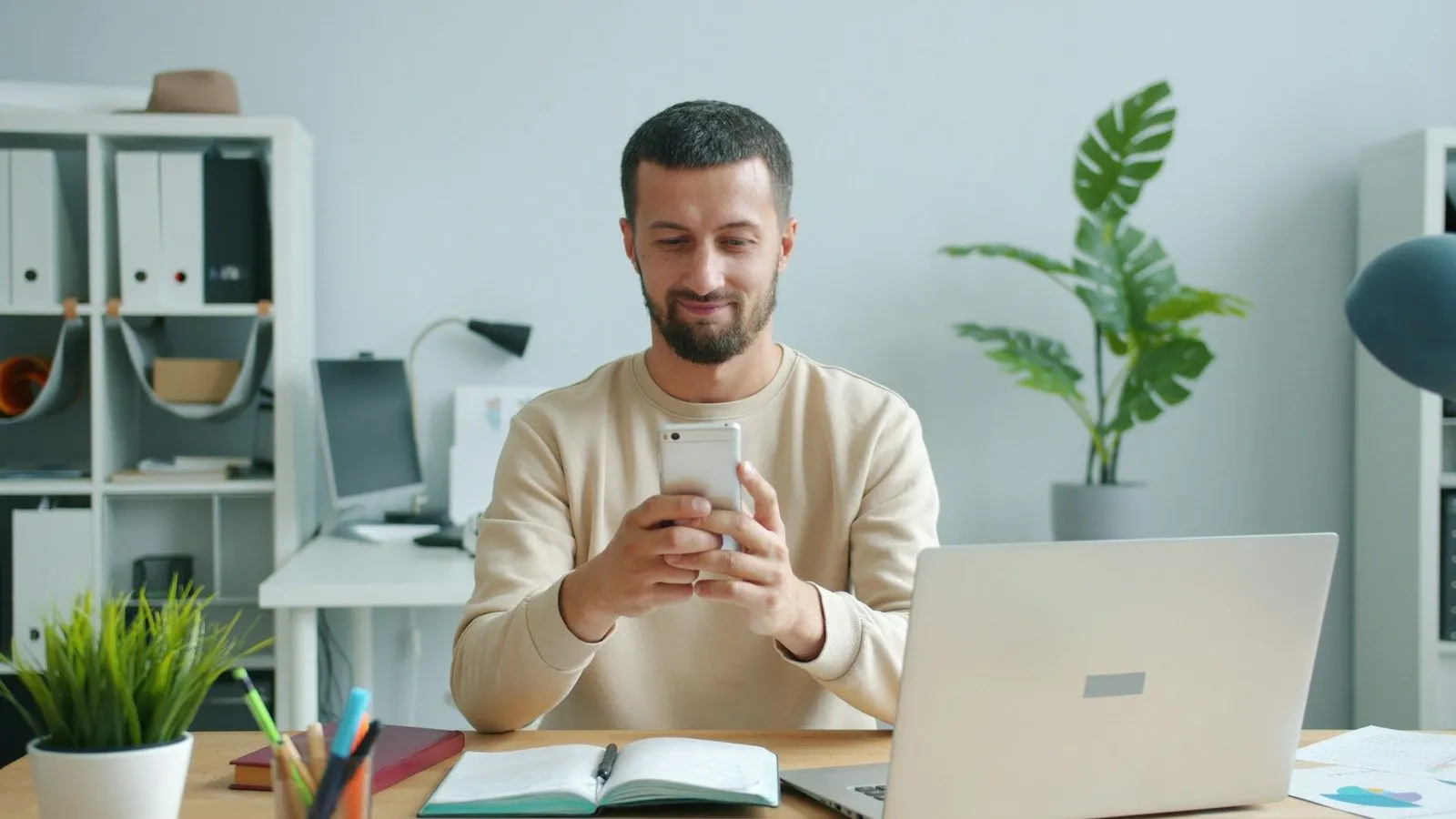 A Flutter architect in a cream sweater smiling at his phone beside a laptop in a bright office.