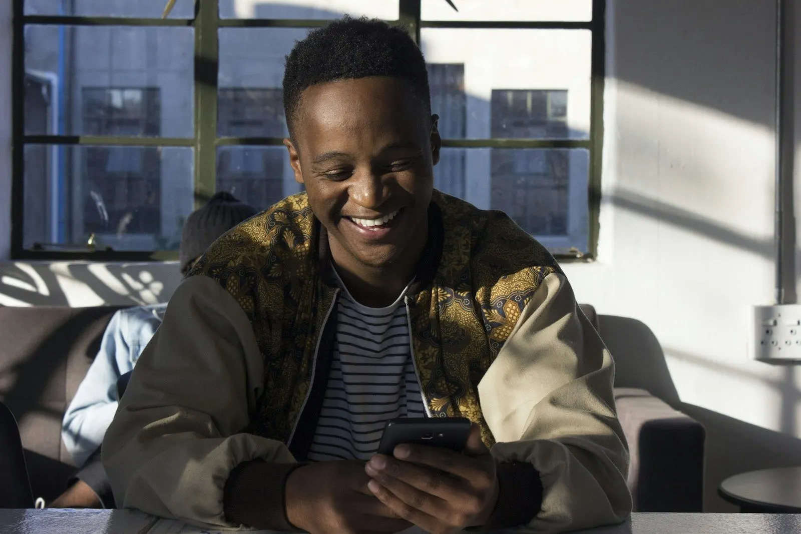 A senior Flutter engineer in a patterned jacket smiling at his phone on a sofa.