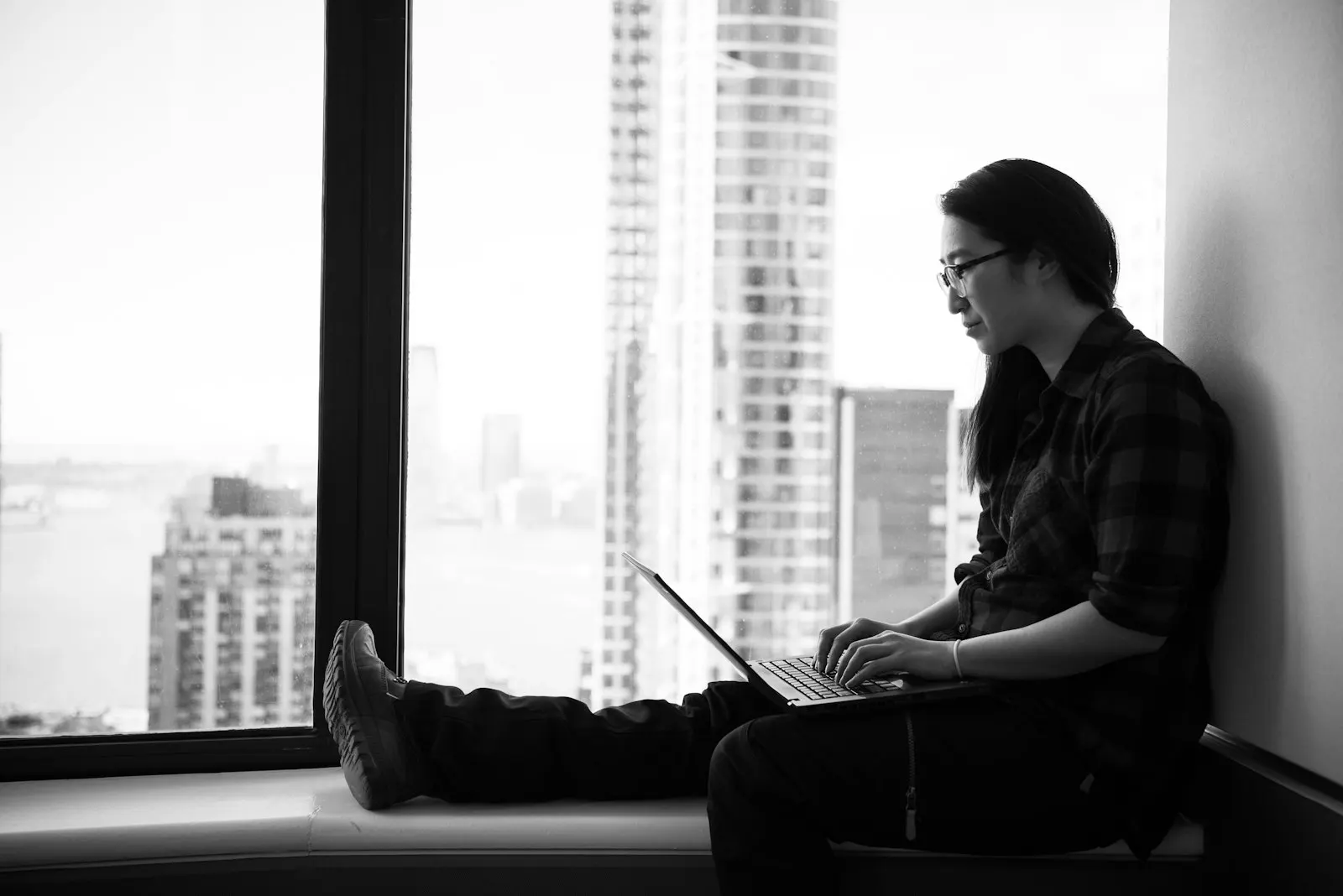 An AI developer working on a laptop on a window ledge high above a city.