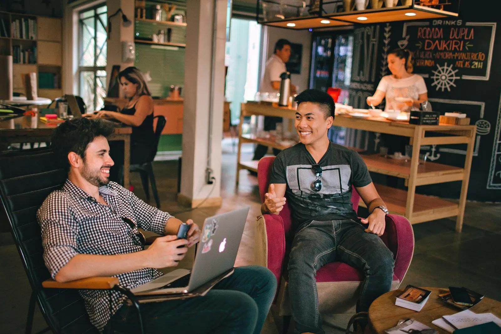 Two data engineers chat over a laptop in a busy cafe-style office.