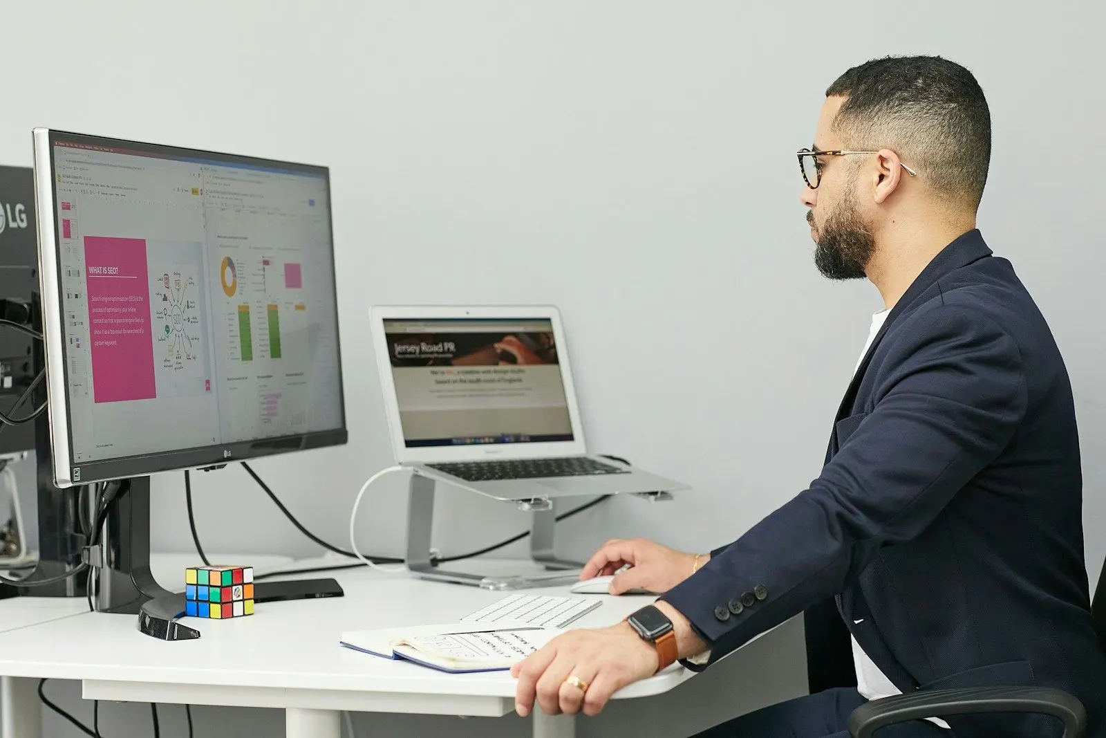 A data engineer in a blazer reviews a warehouse dashboard on a large monitor beside his laptop.