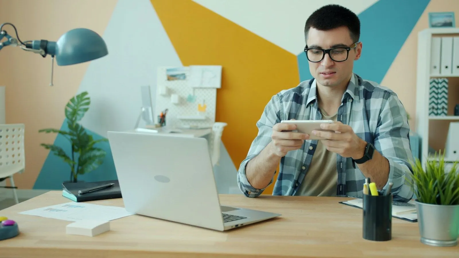 A test automation architect in glasses tests an app on a phone beside his laptop.