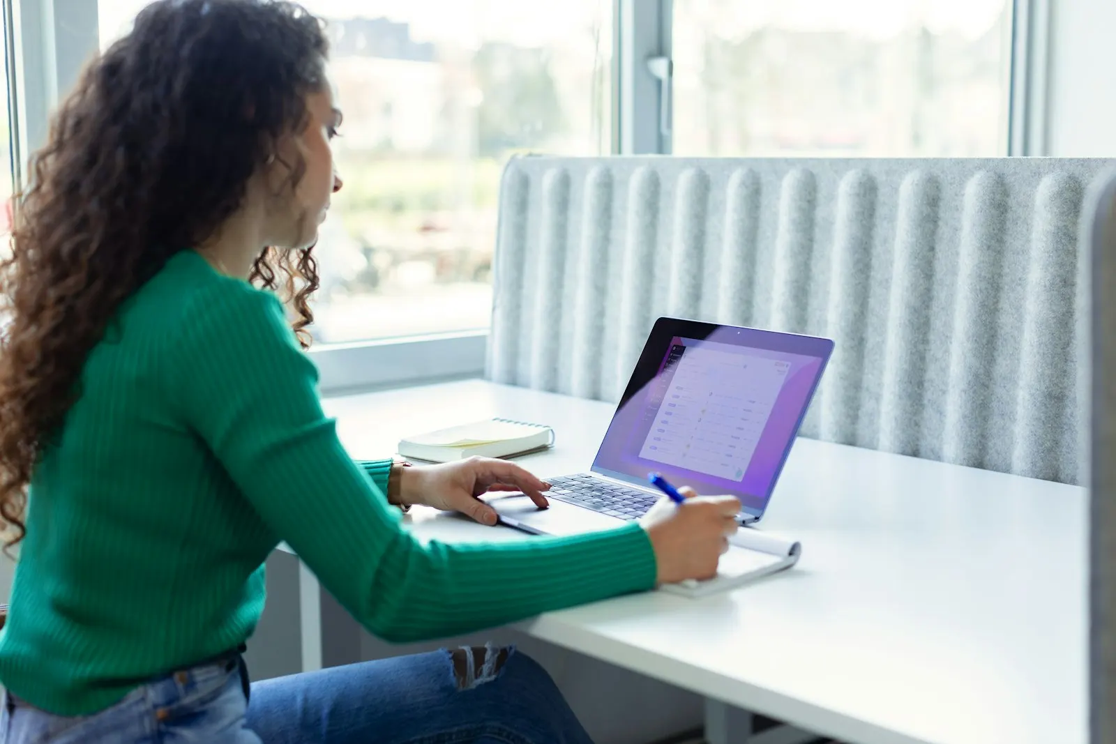 A US onshore QA engineer takes notes beside her laptop at a window desk.