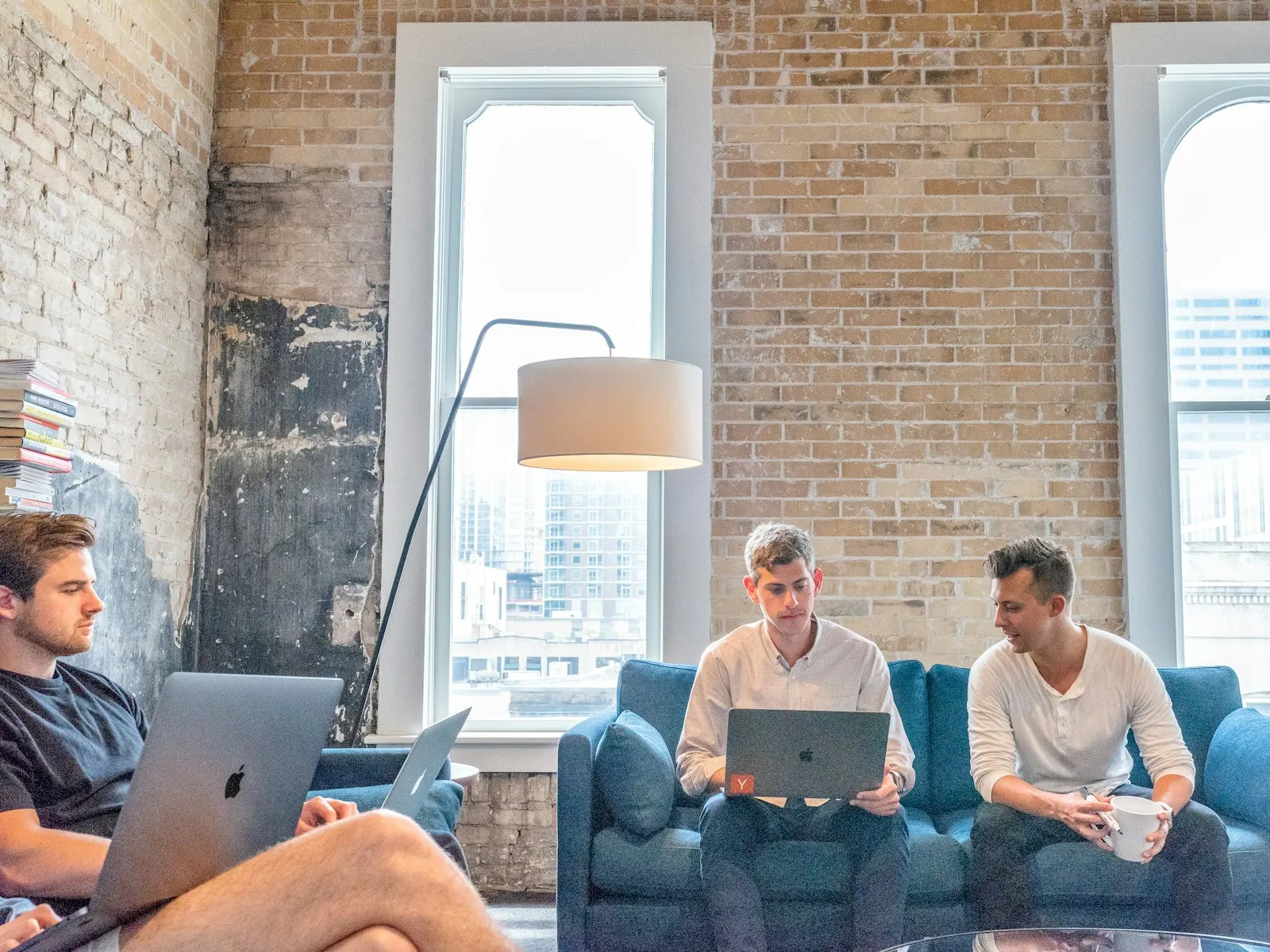 Three onshore developers work on laptops on sofas in a brick-walled office.