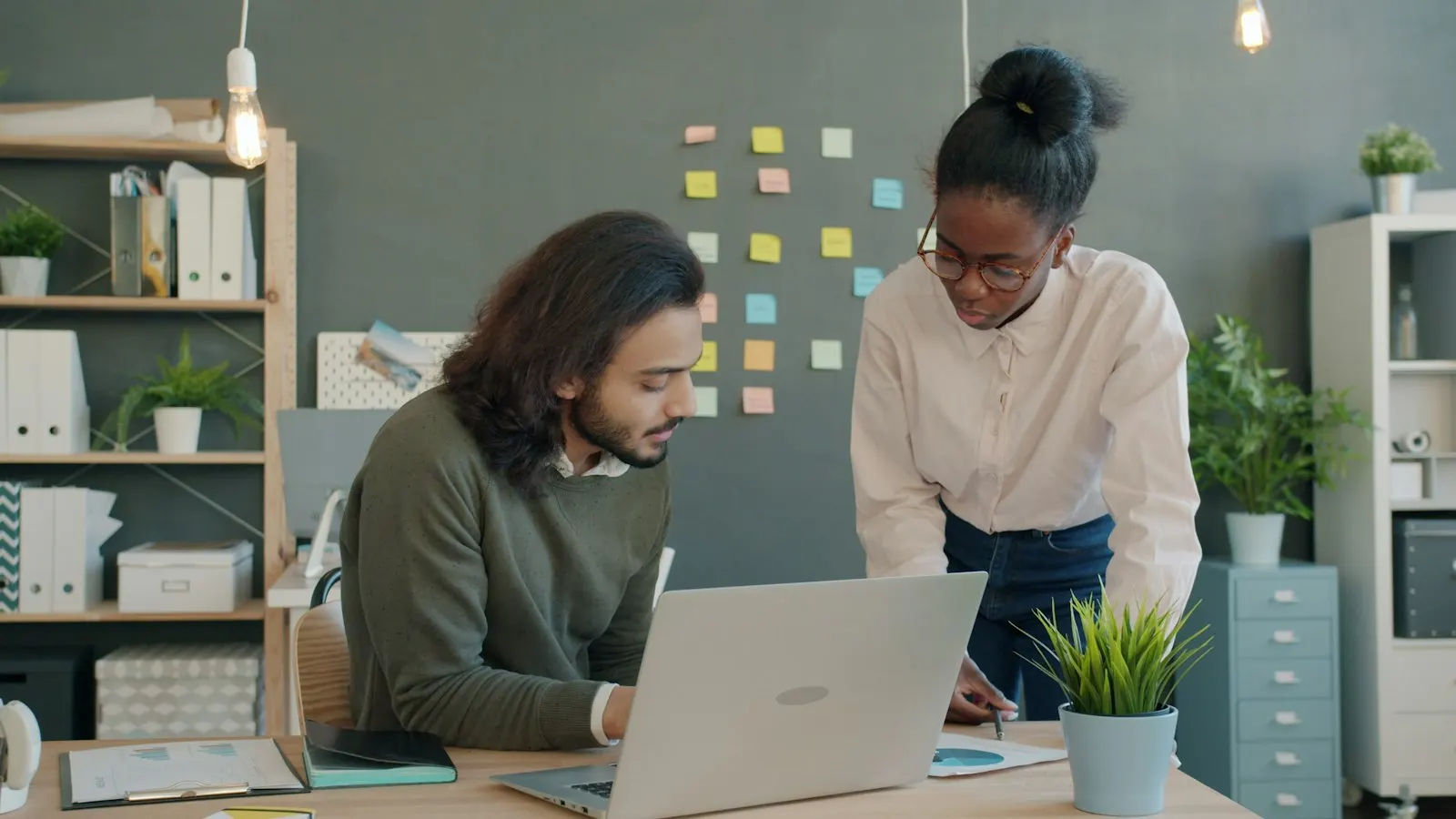 Two colleagues plan together at a desk beside a wall of notes.