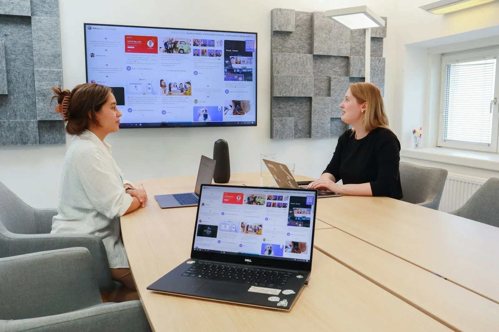 Two QA engineers meet at a conference table with a laptop and a wall screen showing an app.