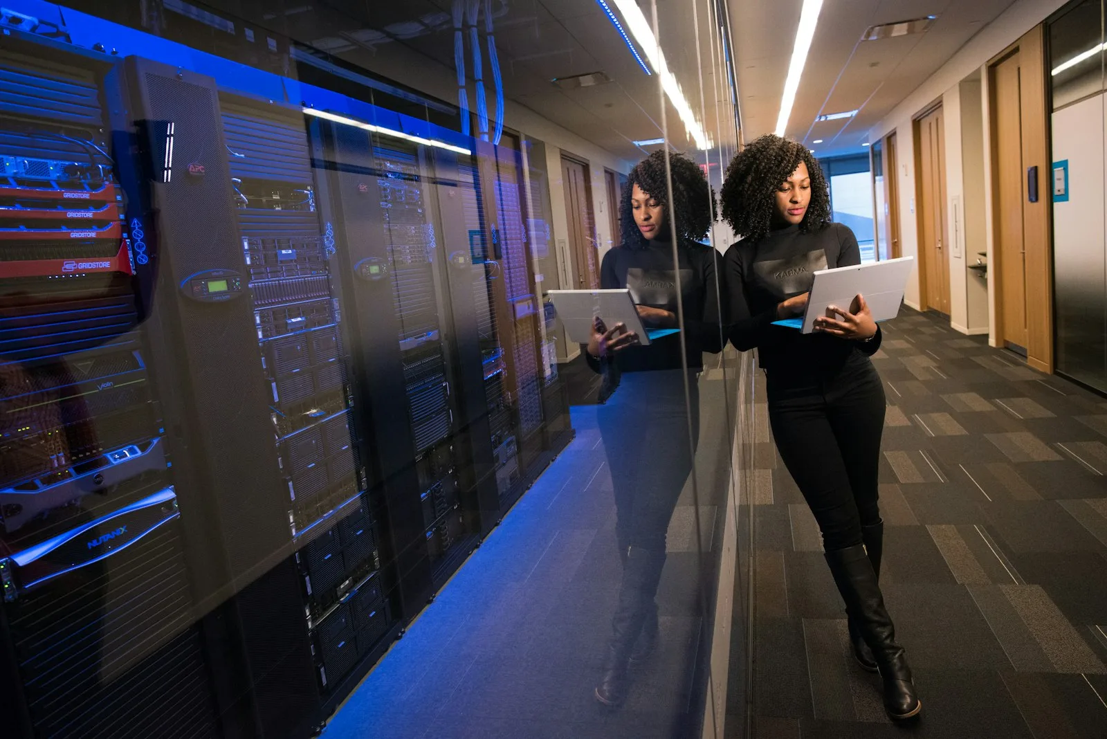 Two hired DevOps Engineers with laptops walk a corridor of server racks lit in blue.