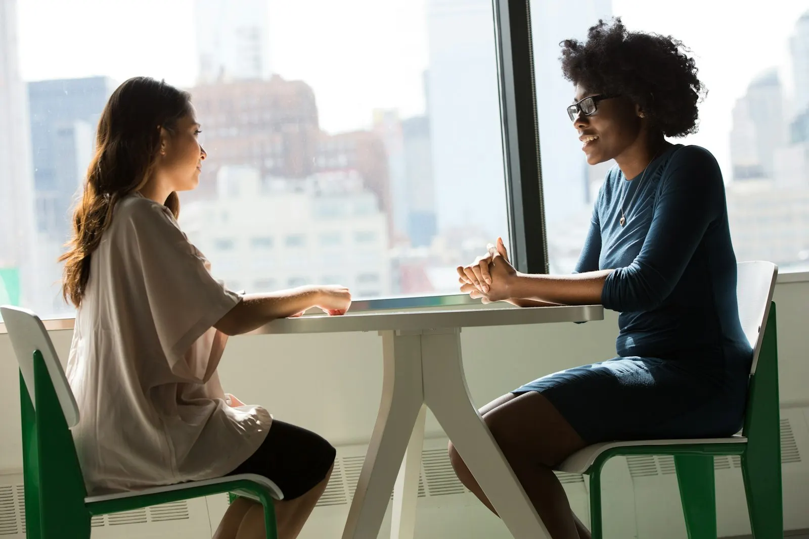 Two colleagues discussing how to hire a specialist developer across a table by a bright window.
